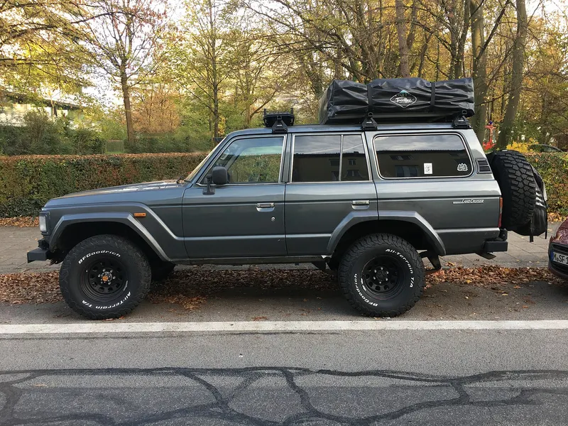 Een grijze terreinwagen staat van opzij gezien geparkeerd langs een straat met herfstbladeren. Op het dak ligt een ingeklapte daktent in een zwarte hoes, en achterop hangt een reservewiel. Op de achtergrond staan een heg en bomen.