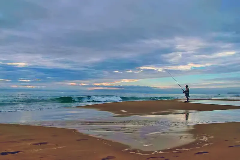 Breed zandstrand bij zonsopkomst onder een bewolkte lucht. In de verte staat een kleine figuur met een hengel op een zandbank tussen plassen zeewater, met brekende golven erachter.