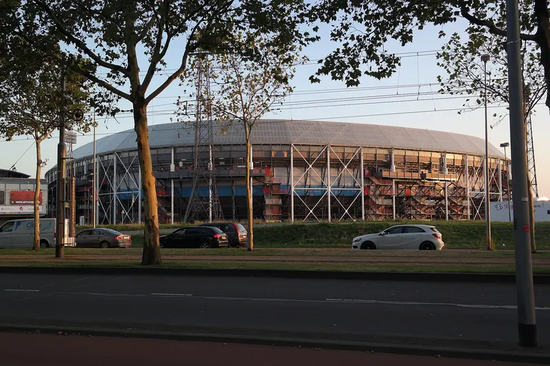 Het ronde stadion De Kuip in Rotterdam bij laag avondlicht, met een zichtbaar stalen skelet en een grijs dak. Op de voorgrond staan bomen, een lege weg en een paar geparkeerde auto's.