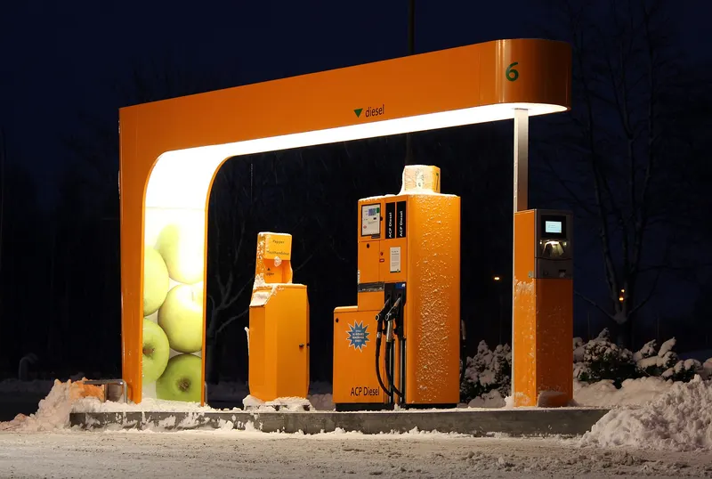 Een verlichte dieselpomp met oranje overkapping bij een tankstation, 's nachts in de sneeuw. Naast de pomp staat een betaalautomaat. Er zijn geen mensen te zien.