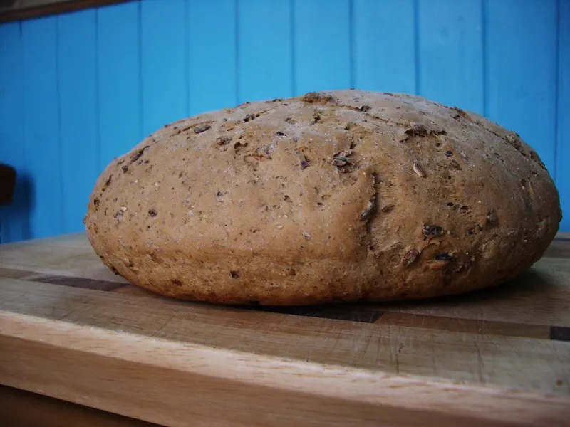 Een rond, heel bruin brood met zaden en een stevige korst ligt op een houten snijplank, voor een lichtblauwe houten wand.