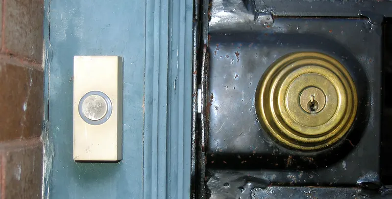 Close-up van een voordeur: links een lichte deurbel met ronde knop op een blauwgroen geverfd kozijn naast bakstenen, rechts een messing cilinderslot op een donkere metalen deur.