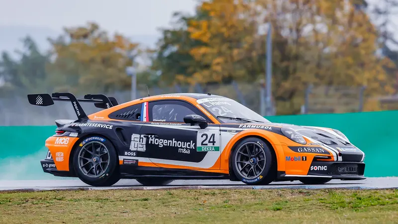 Een oranje-zwarte Porsche 911 met startnummer 24 rijdt van opzij gezien over een nat circuit, met een groene vangrail en herfstbomen op de achtergrond.
