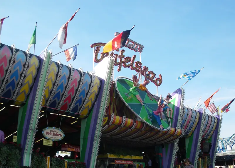 De kleurrijke gevel van de kermisattractie Teufelsrad met de naam in lichtletters, een beschilderde draaischijf met figuren en wapperende vlaggen tegen een blauwe lucht.