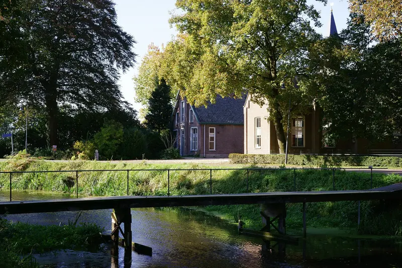 Een houten loopbrug over een smalle singel met groene oevers. Erachter staan bakstenen huizen tussen hoge bomen, met rechtsboven de spits van een kerktoren.